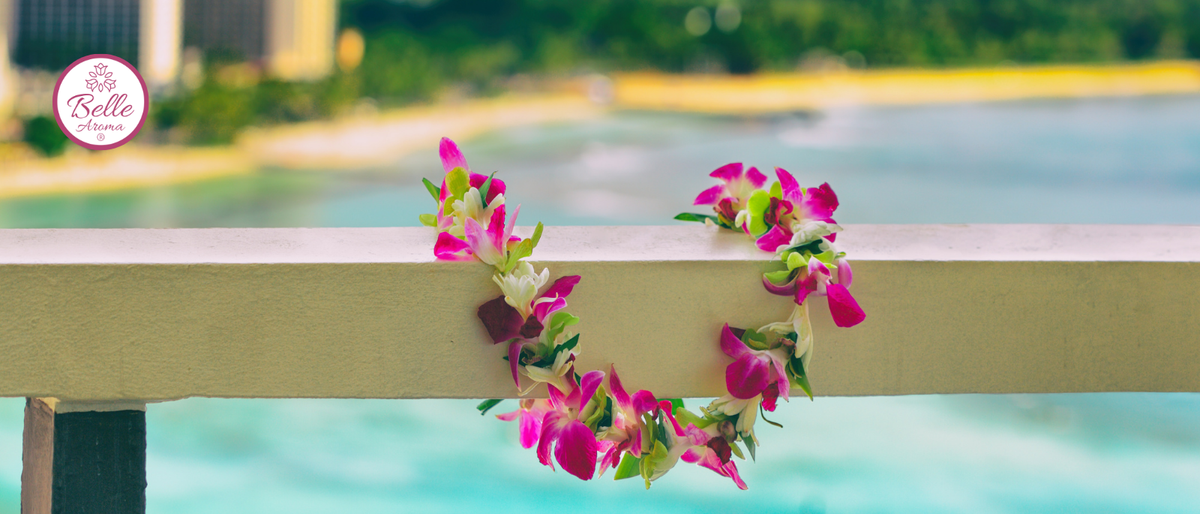 Pink and white orchid lei draped over a hotel balcony railing, with turquoise water and a tropical shoreline in the background. Belle Aroma logo appears in the upper left.