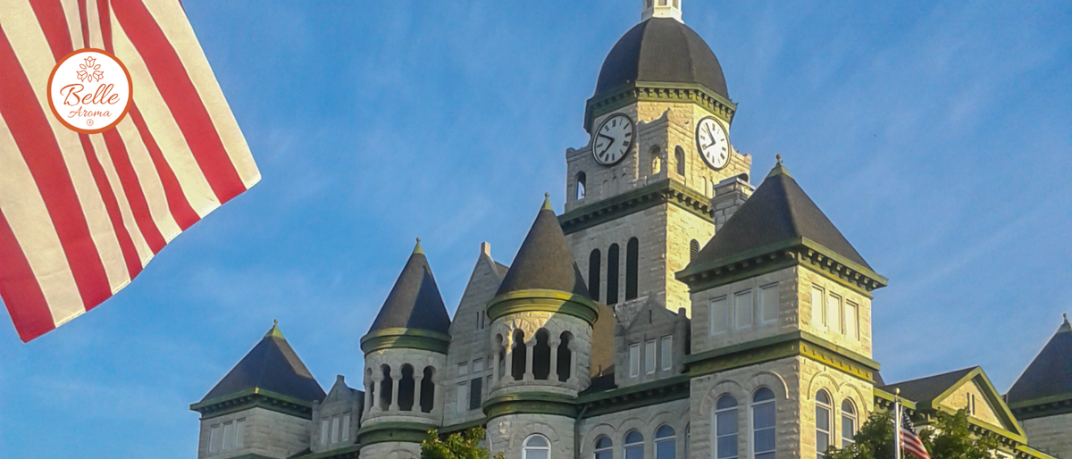 American flag waving beside a historic stone courthouse with a clock tower under a bright blue sky, featuring the Belle Aroma logo.